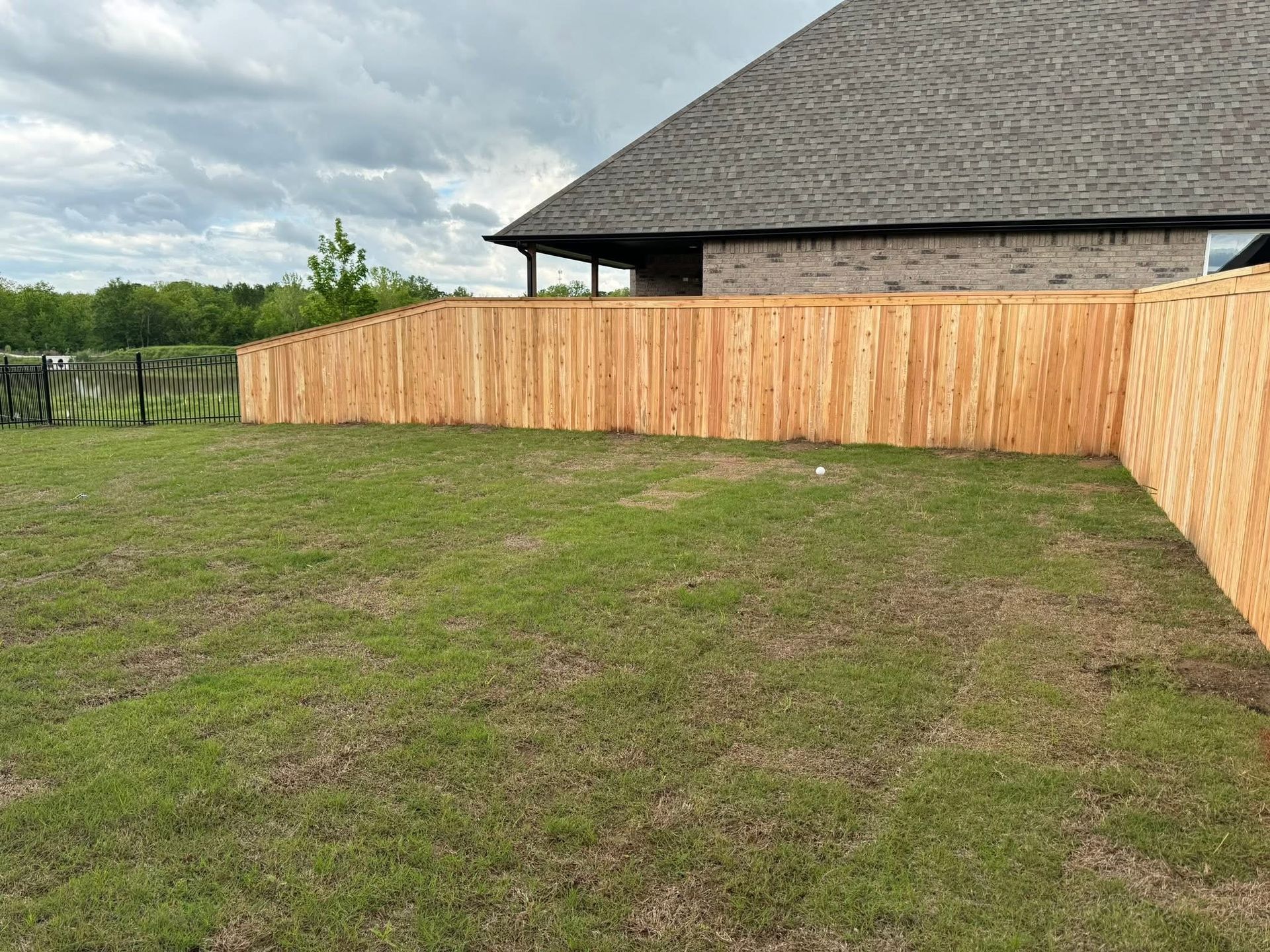 Backyard with patchy grass and a long wooden fence beside a brick house under cloudy sky