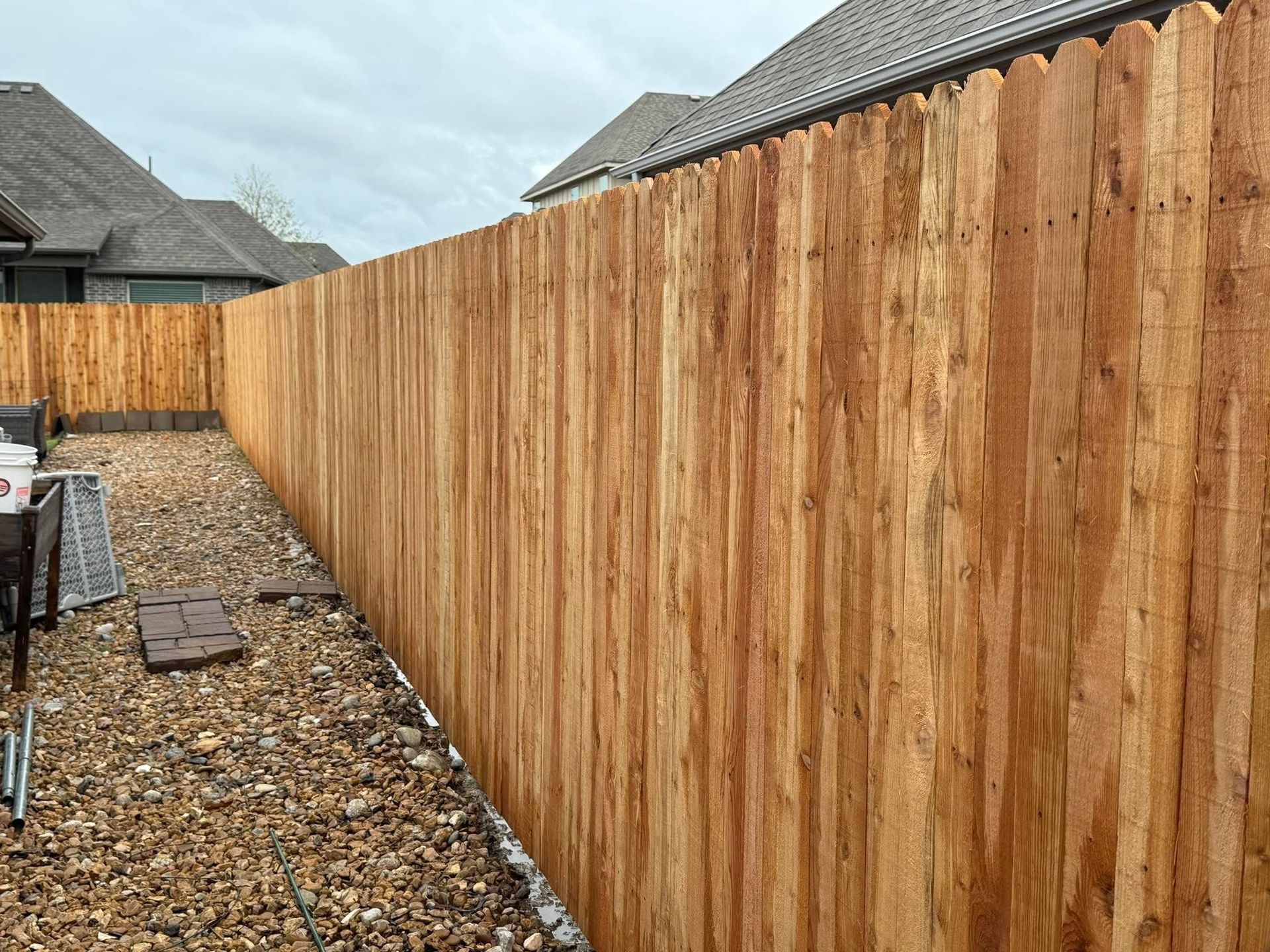 Wooden privacy fence lining a gravel backyard beside neighboring houses under a cloudy sky