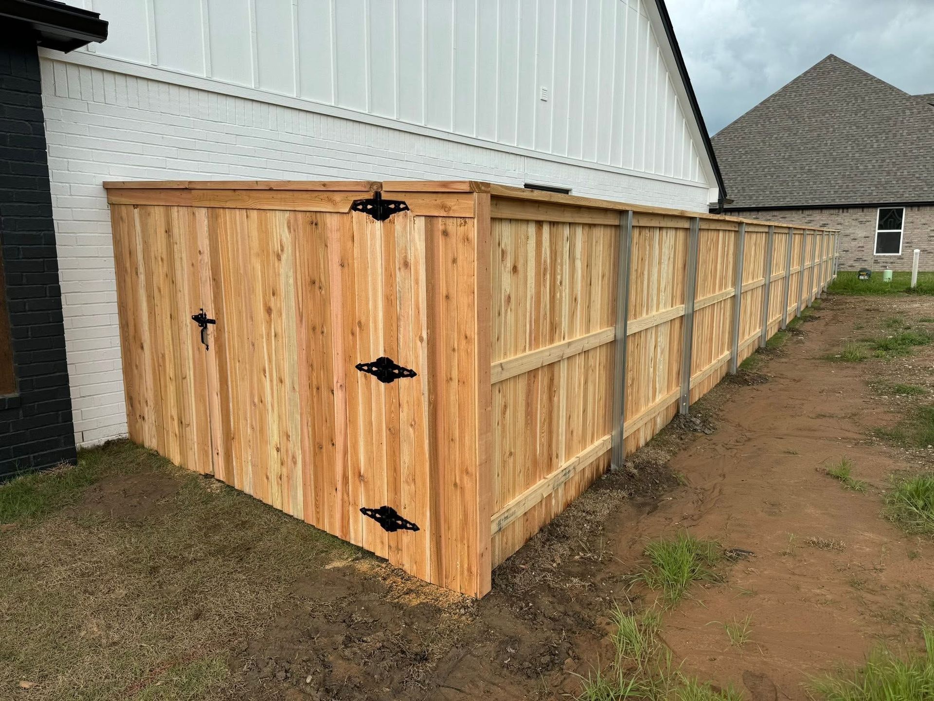 Wooden privacy fence attached to a white house, with black hinges and a dirt yard alongside it.
