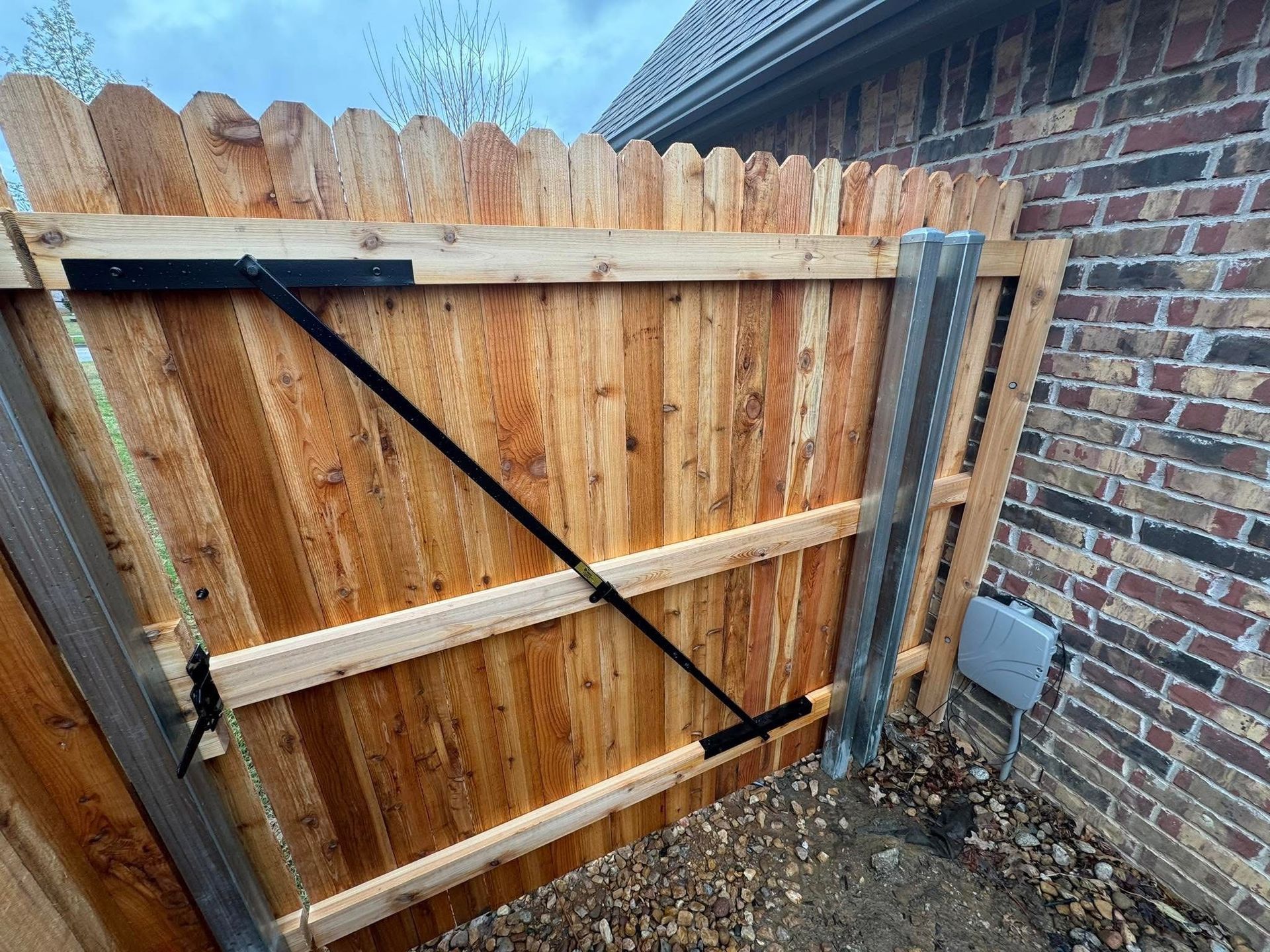 Wooden backyard gate with black diagonal brace, beside a brick wall and gravel ground.