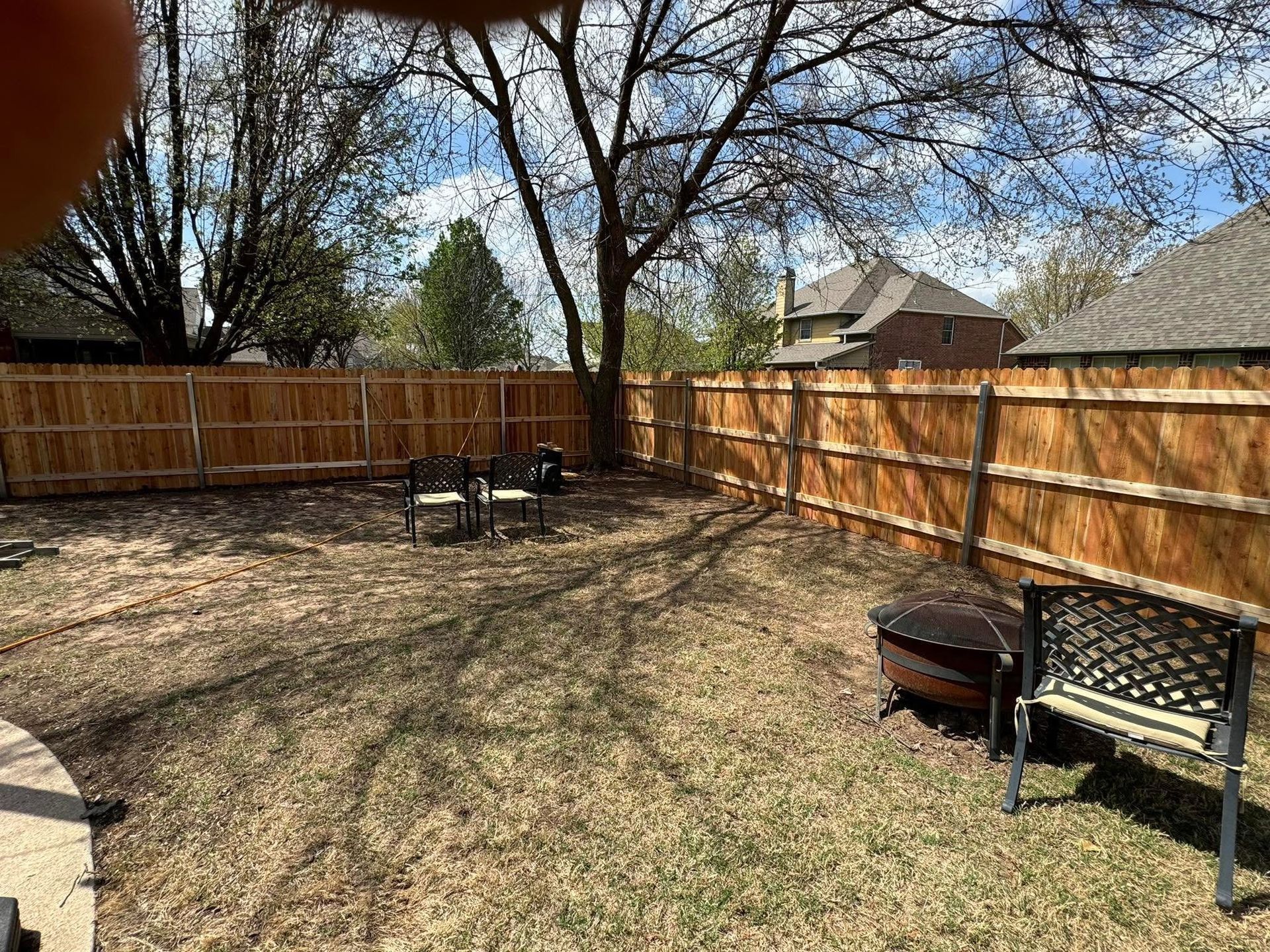 Backyard with wooden fence, leafless trees, patio chairs, and a fire pit in a sunny yard