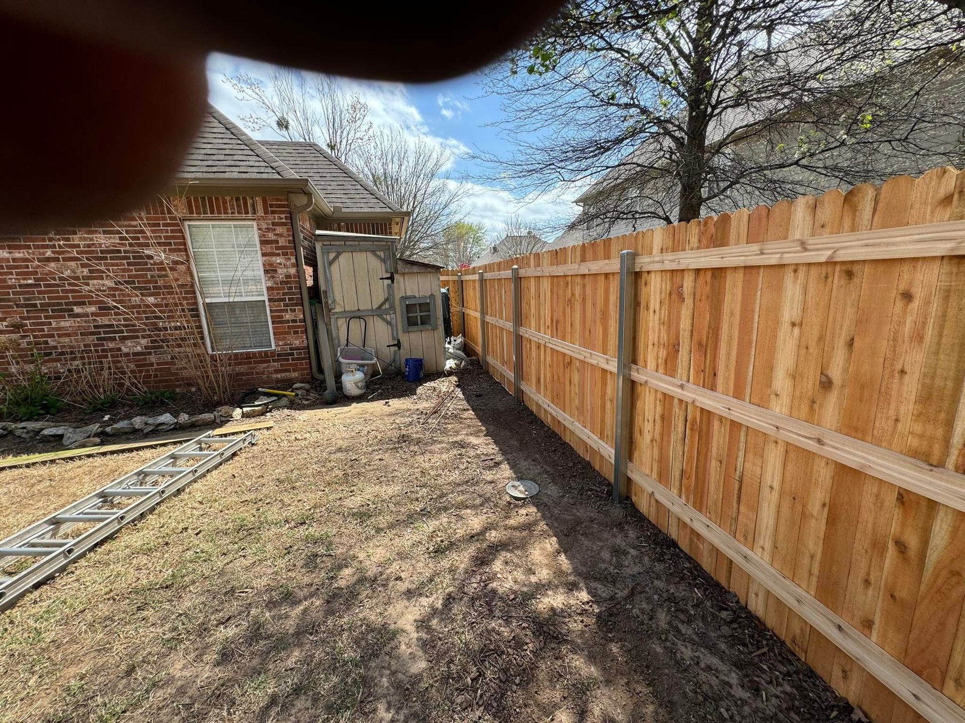 Narrow backyard with wooden fence, brick house, and patchy grass under a blue sky.