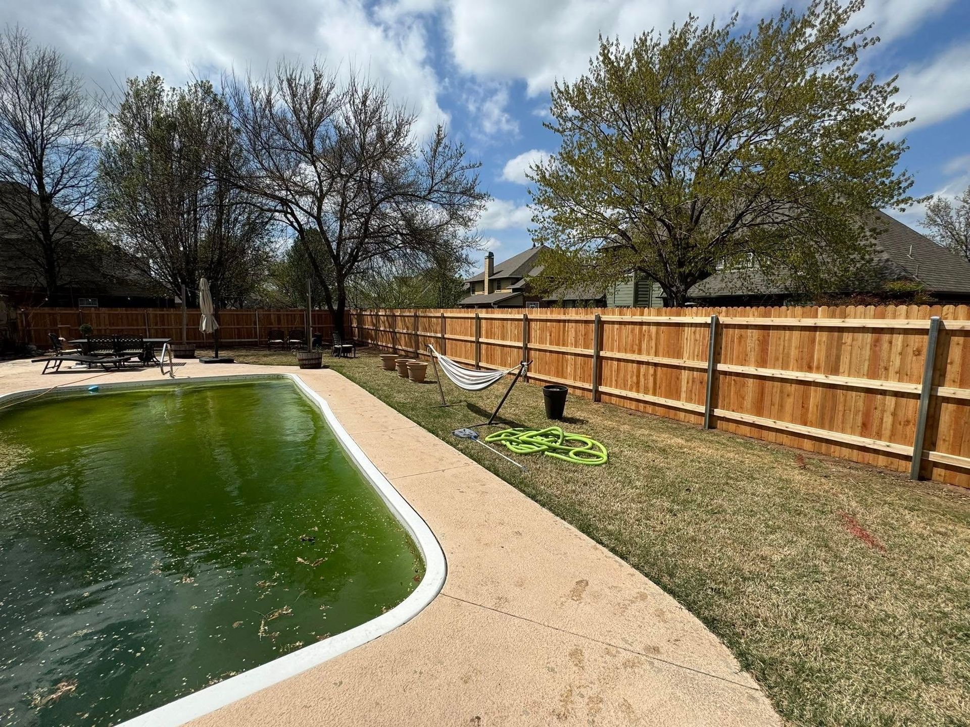Backyard pool with green water, beige deck, wooden fence, trees, and cloudy sky
