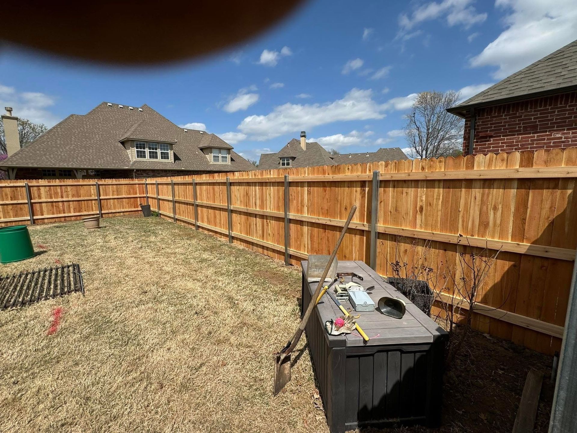 Backyard with wooden fence, stone patio, trash bin, and suburban houses under a blue sky