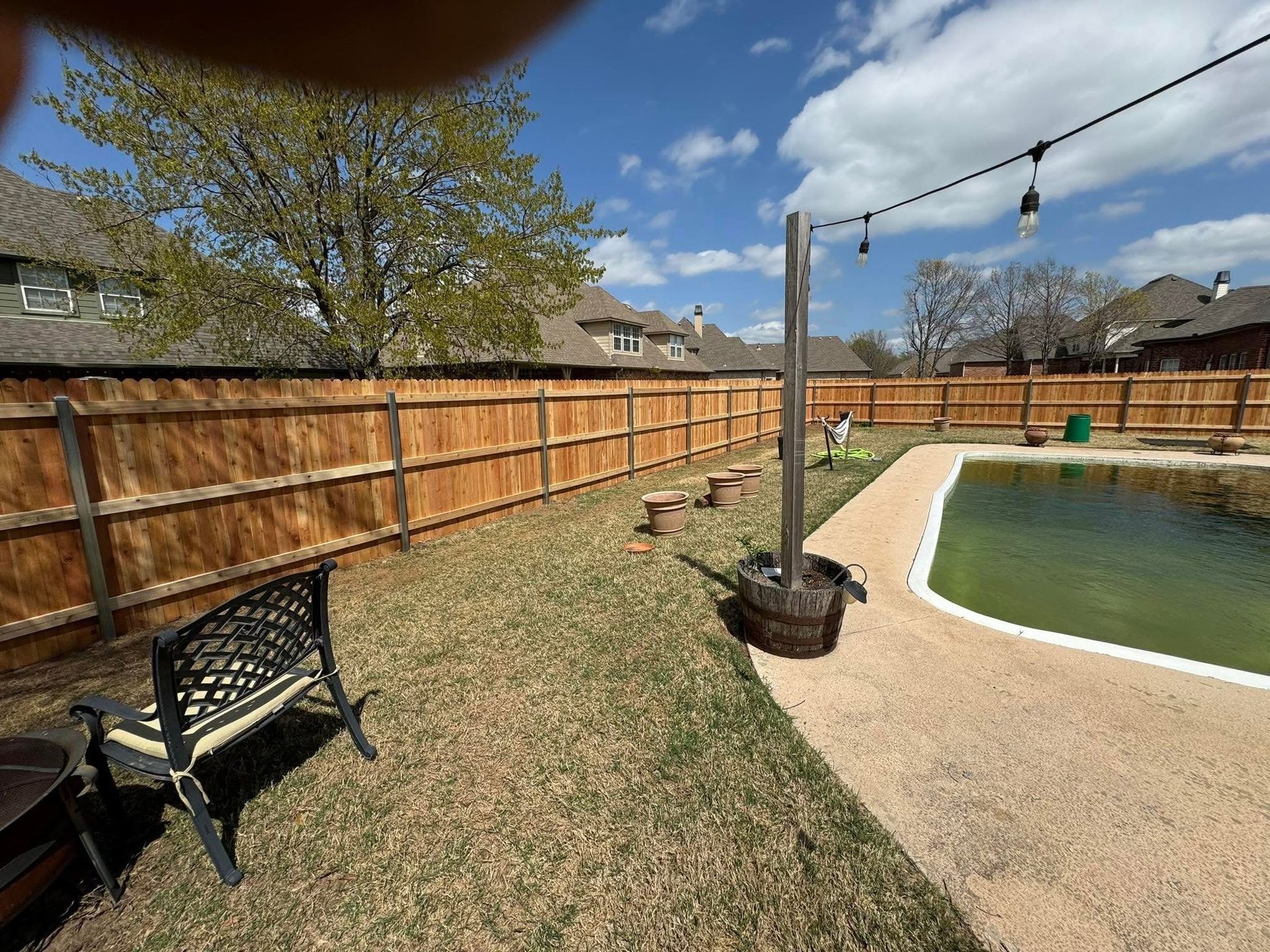 Backyard patio with a pool, wooden fence, bench, and string lights under a blue sky