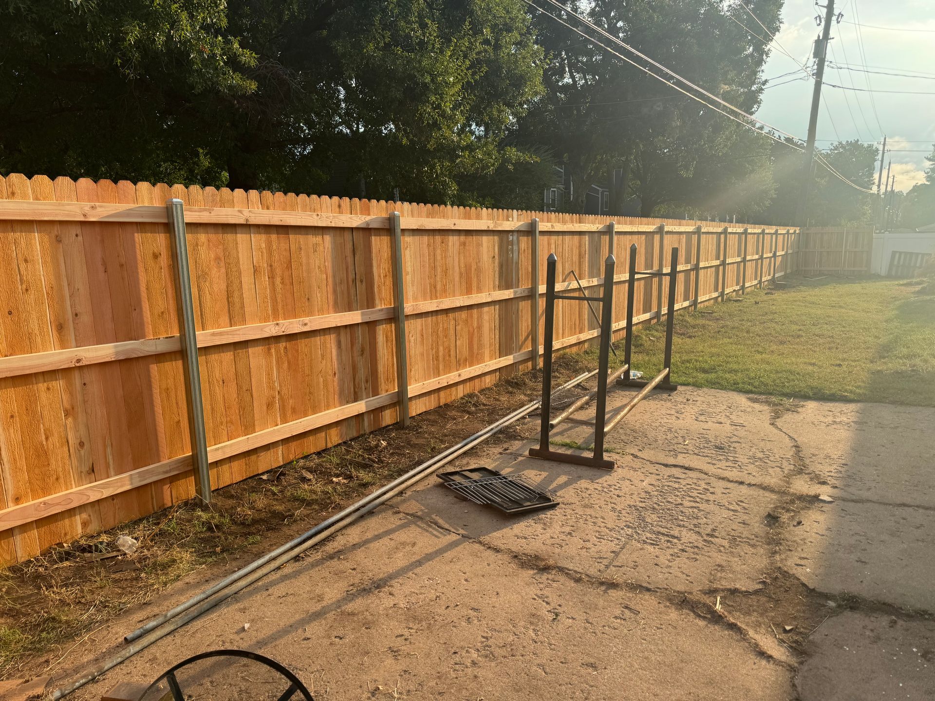 A long wooden fence beside a cracked driveway with a metal rack and garden tools in sunlight.