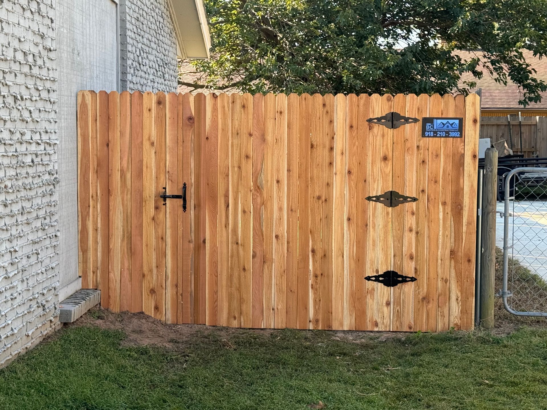 Wooden privacy gate with black hinges beside a white brick house and grassy yard