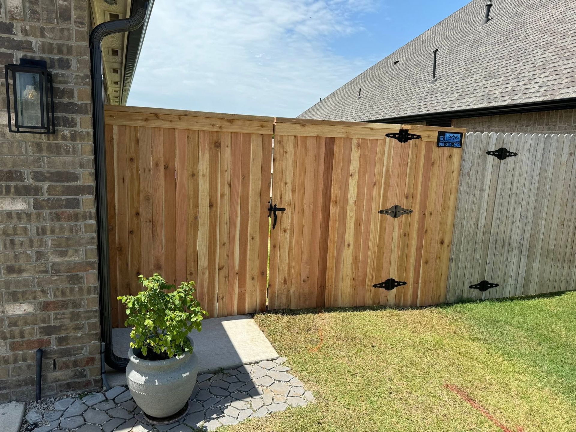 Wooden fence gate beside a brick house, with black hinges and a potted plant on a patio.