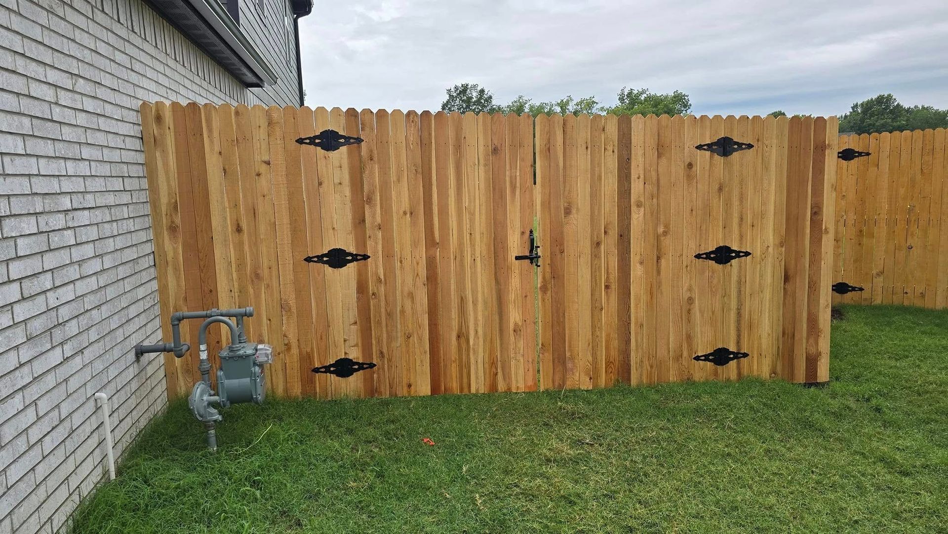 Wooden gate with black hinges beside a brick house, opening to a grassy backyard under an overcast sky.