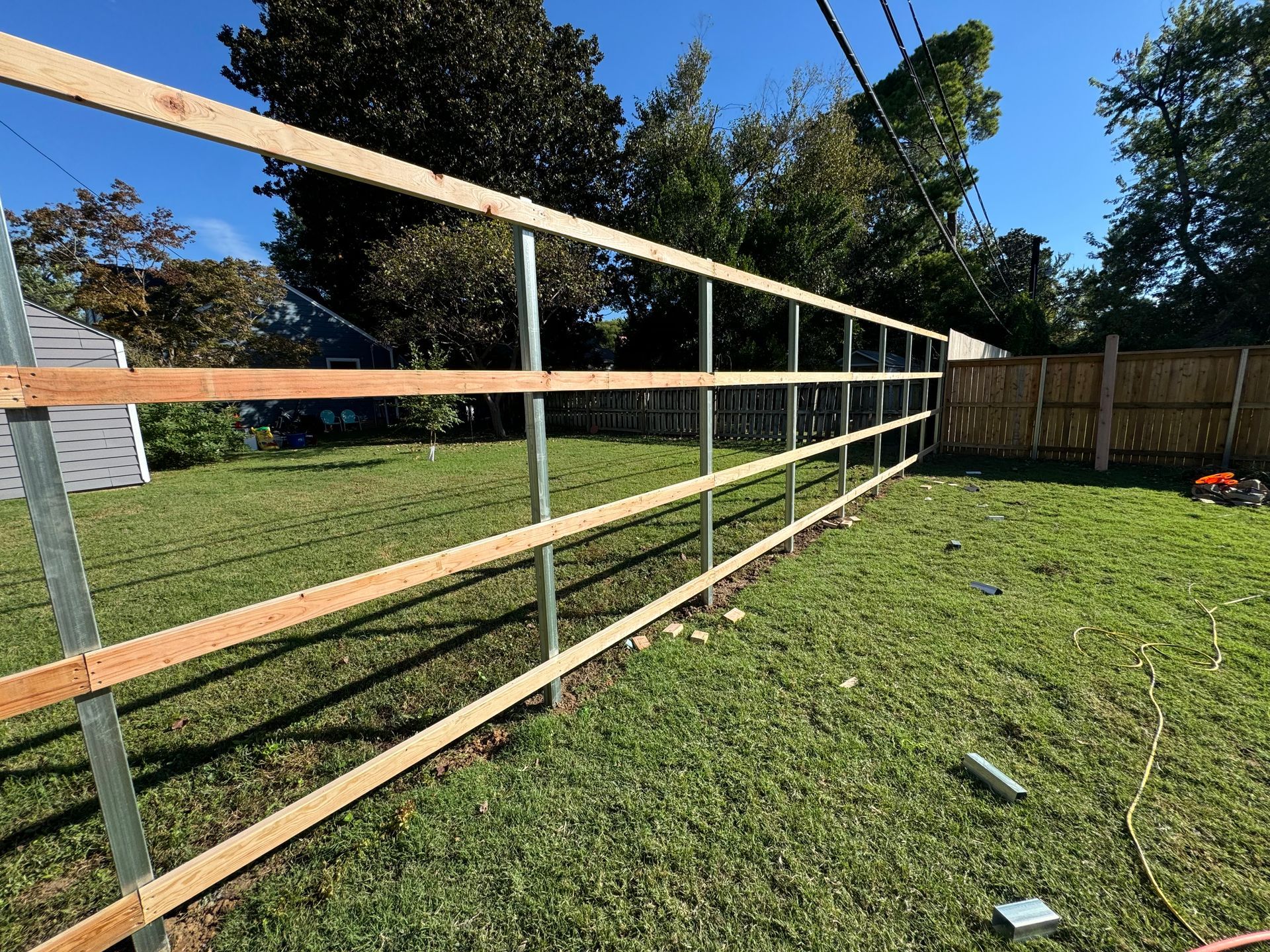 Wooden fence rails and posts along a sunny grassy backyard with trees and a wood privacy fence