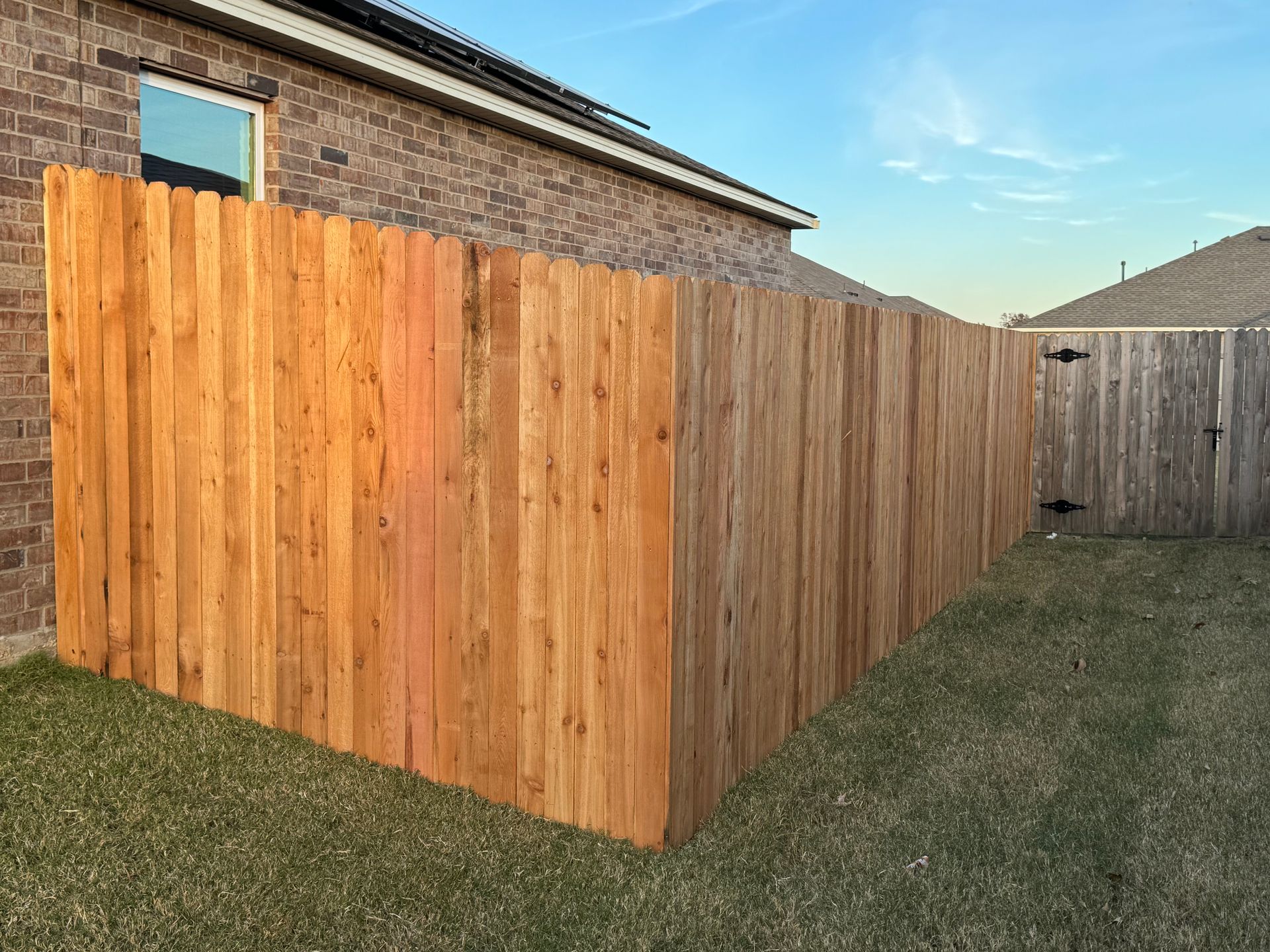 Freshly stained wooden fence enclosing a backyard beside a brick house and grassy lawn