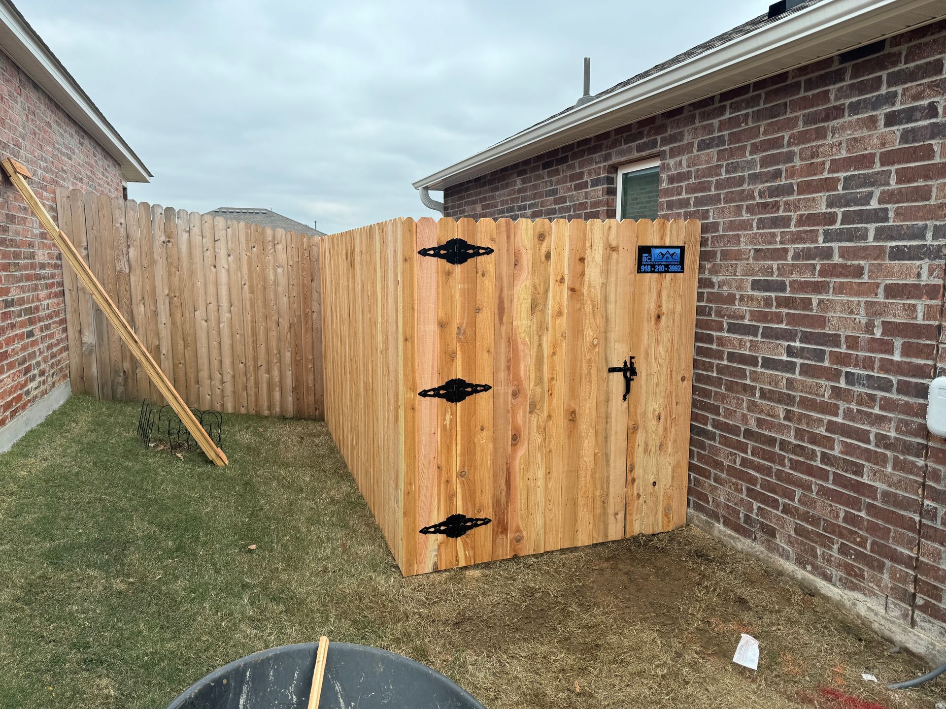 Wooden privacy enclosure beside a brick house, with a gate and black hinges in a narrow side yard.