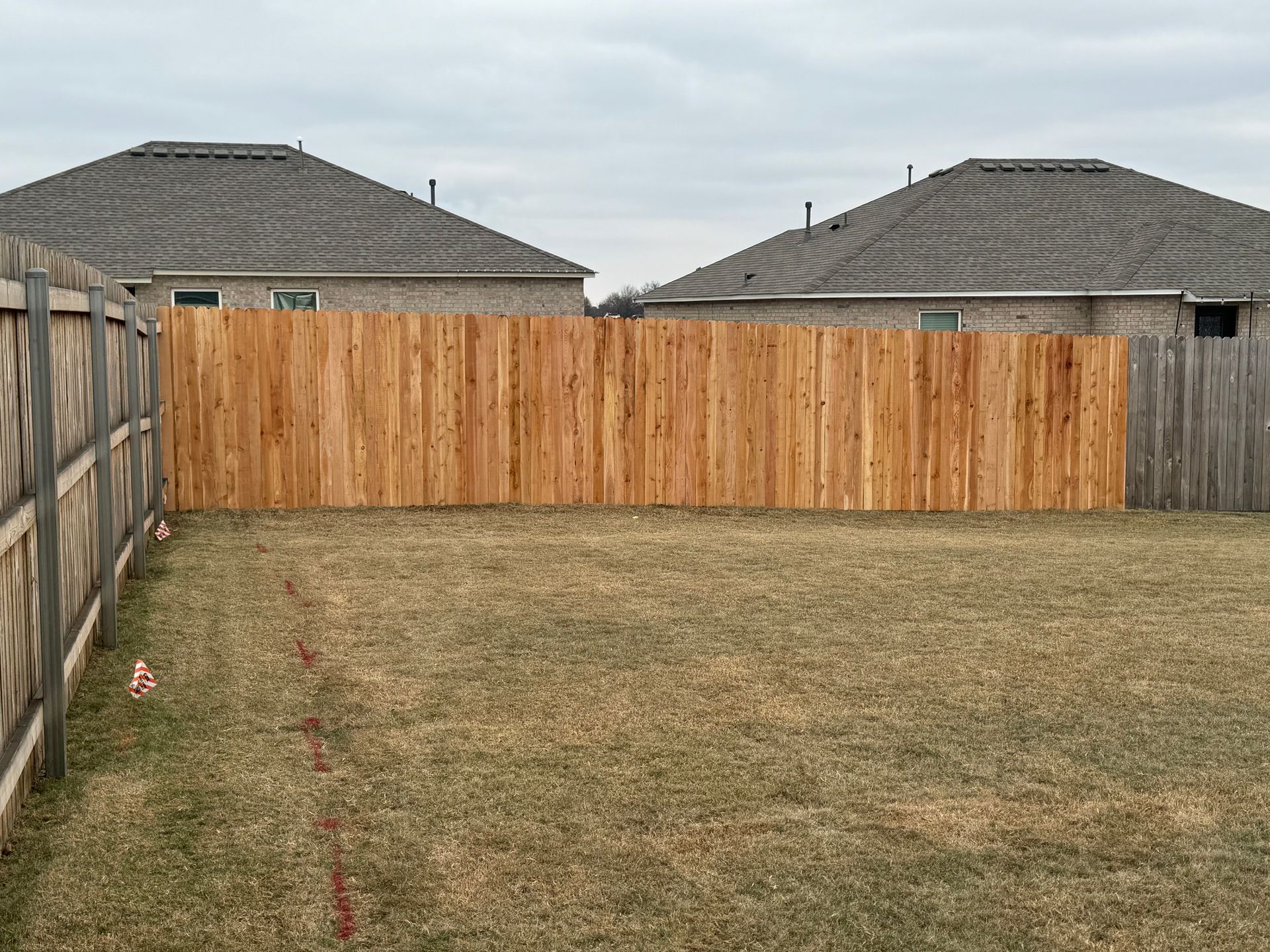 Backyard with a wooden privacy fence, patchy grass, and neighboring house roofs visible.