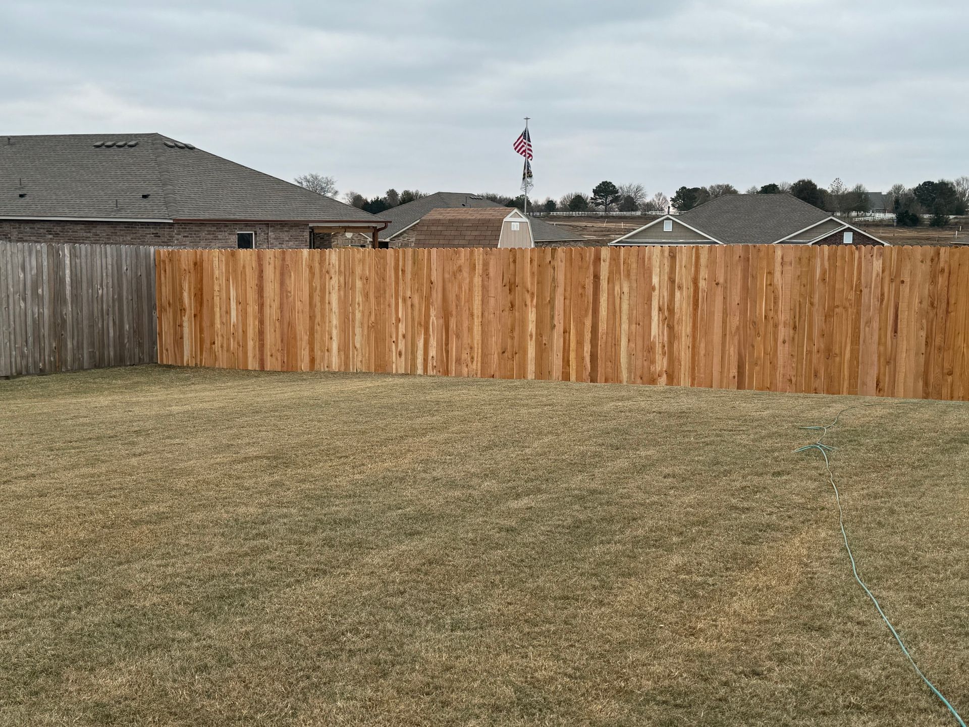 Suburban backyard with dry grass, a wooden fence, and rooftops under an overcast sky