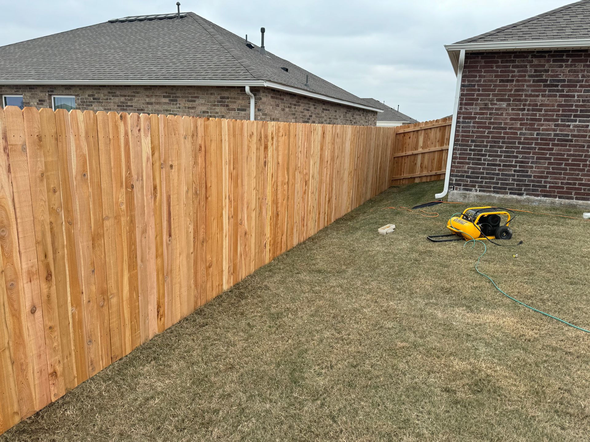 Wooden privacy fence along a grassy side yard between brick houses, with a yellow mower on the ground.