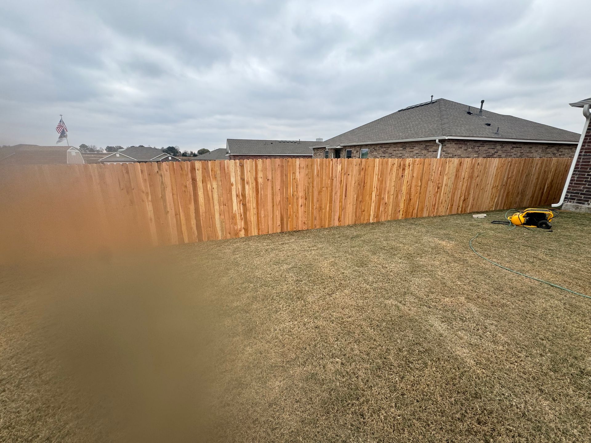 Backyard with wooden fence, dry grass, cloudy sky, and a small mower near the right edge
