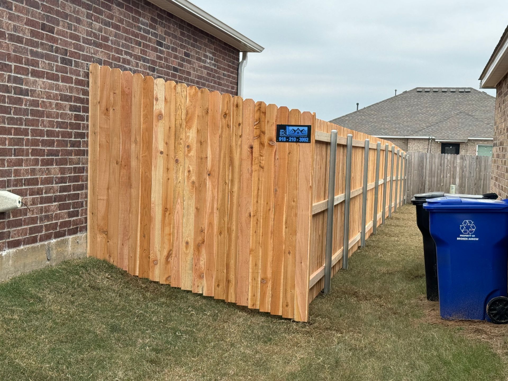 Wooden privacy fence beside a brick house, with blue recycling bins along a narrow side yard.