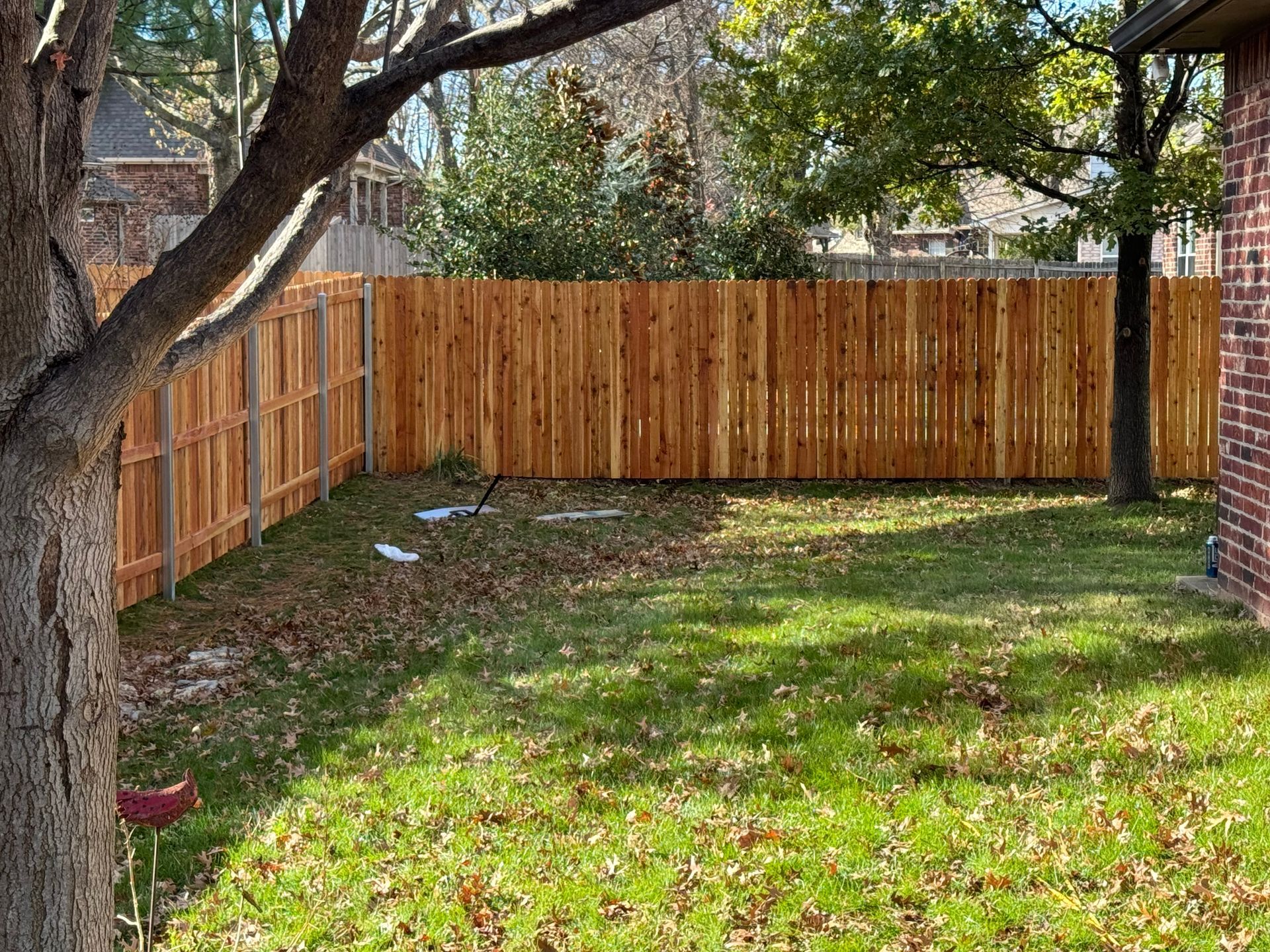 Small backyard with green grass, wooden fence, large tree, and a brick house wall on the right.