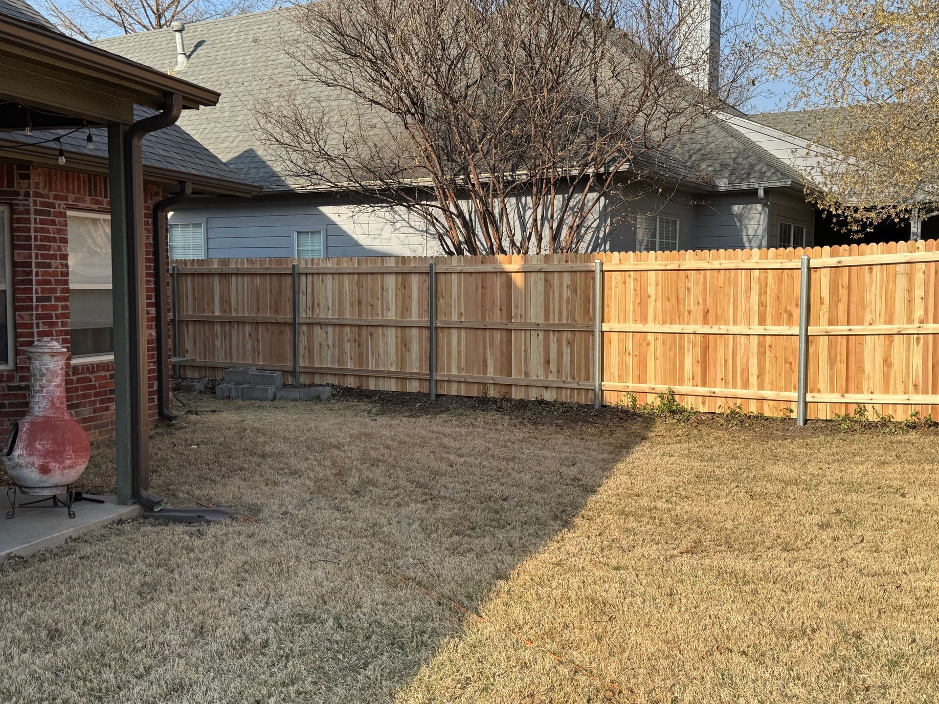 Backyard with dry grass, wooden fence, and part of a brick house under bright sunlight