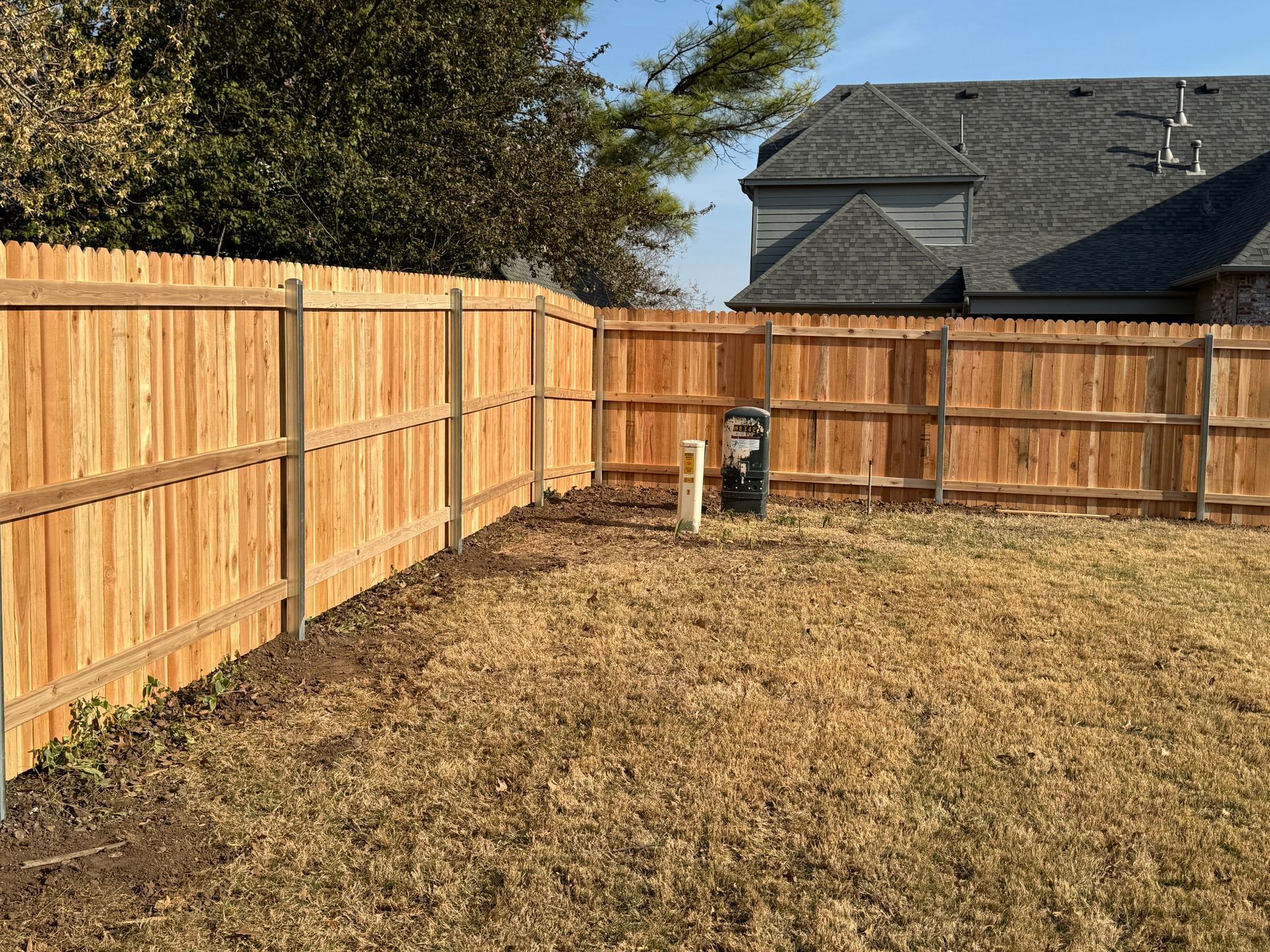 Backyard with a wooden fence, dry grass, and utility pipes near a house.