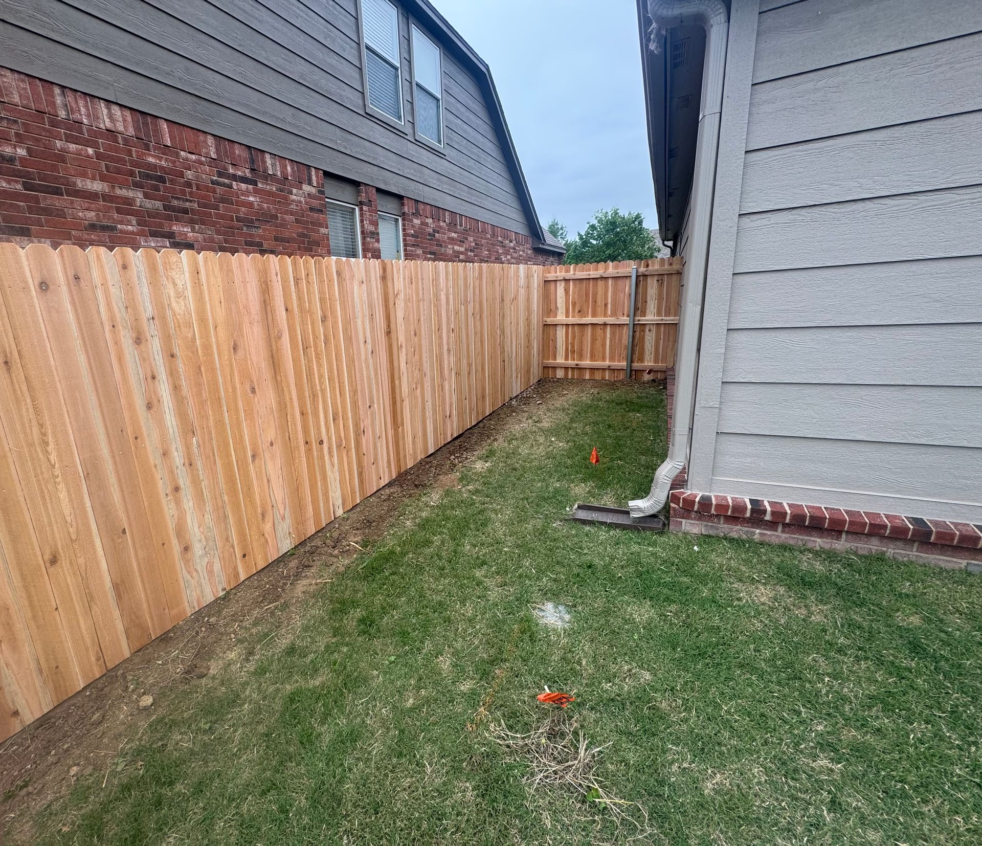 Narrow grassy side yard between two houses with wooden fence and gray siding, with a red marker near the ground