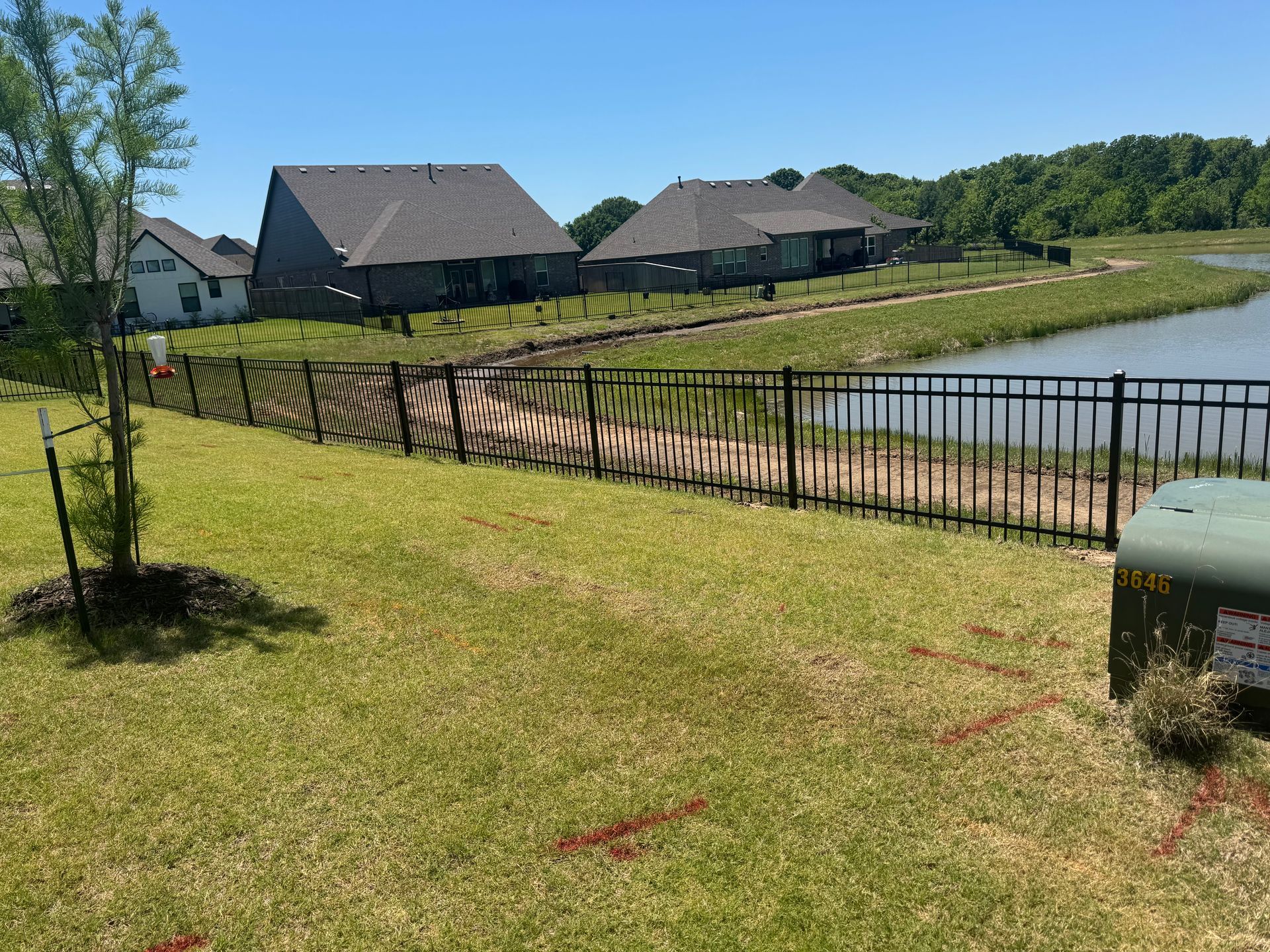 Grassy lakeside yard with black fence, curved path, and dark-roofed buildings under a clear blue sky