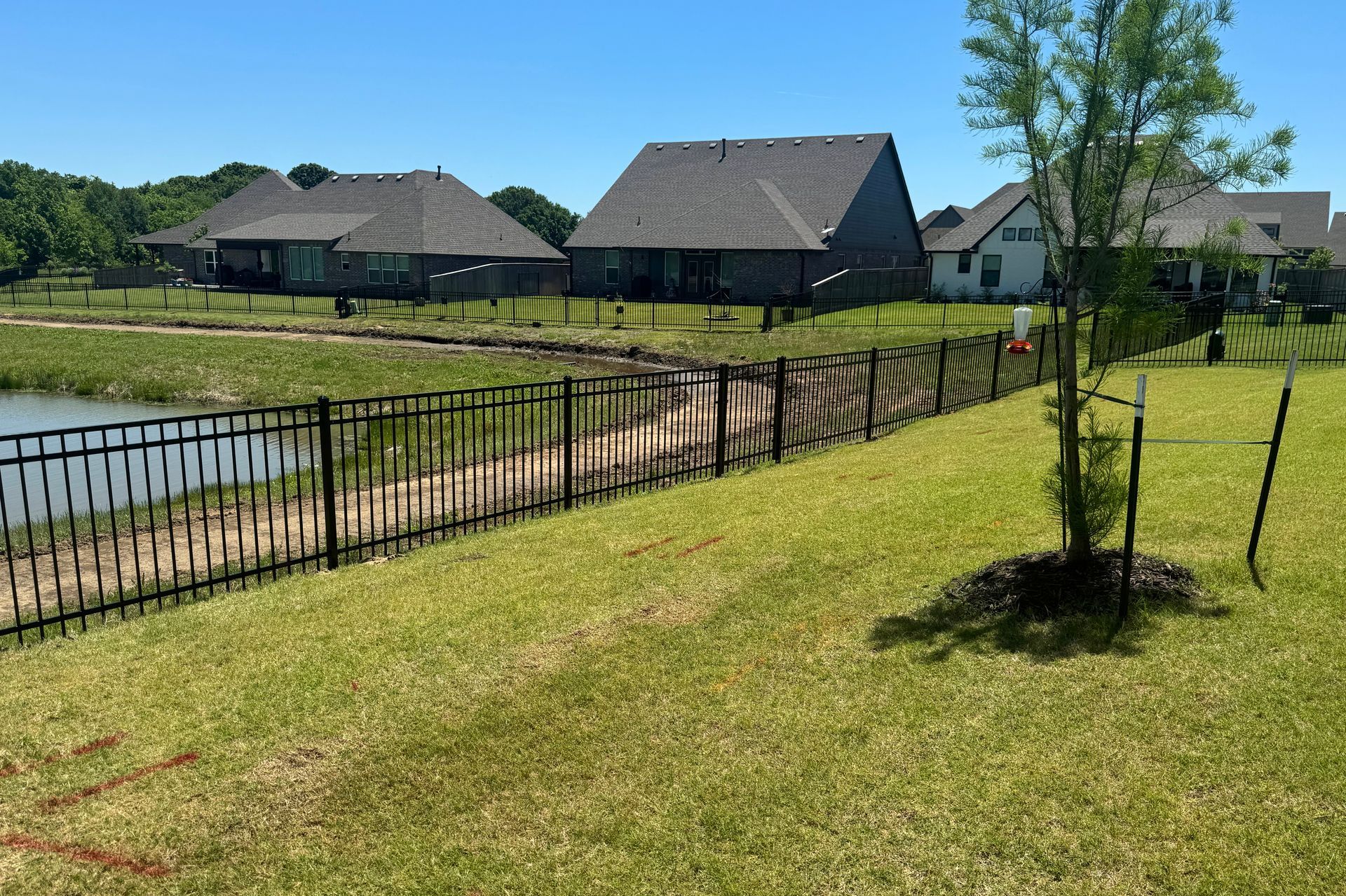 Grassy riverside path with black fence, small trees, and rustic houses across the water
