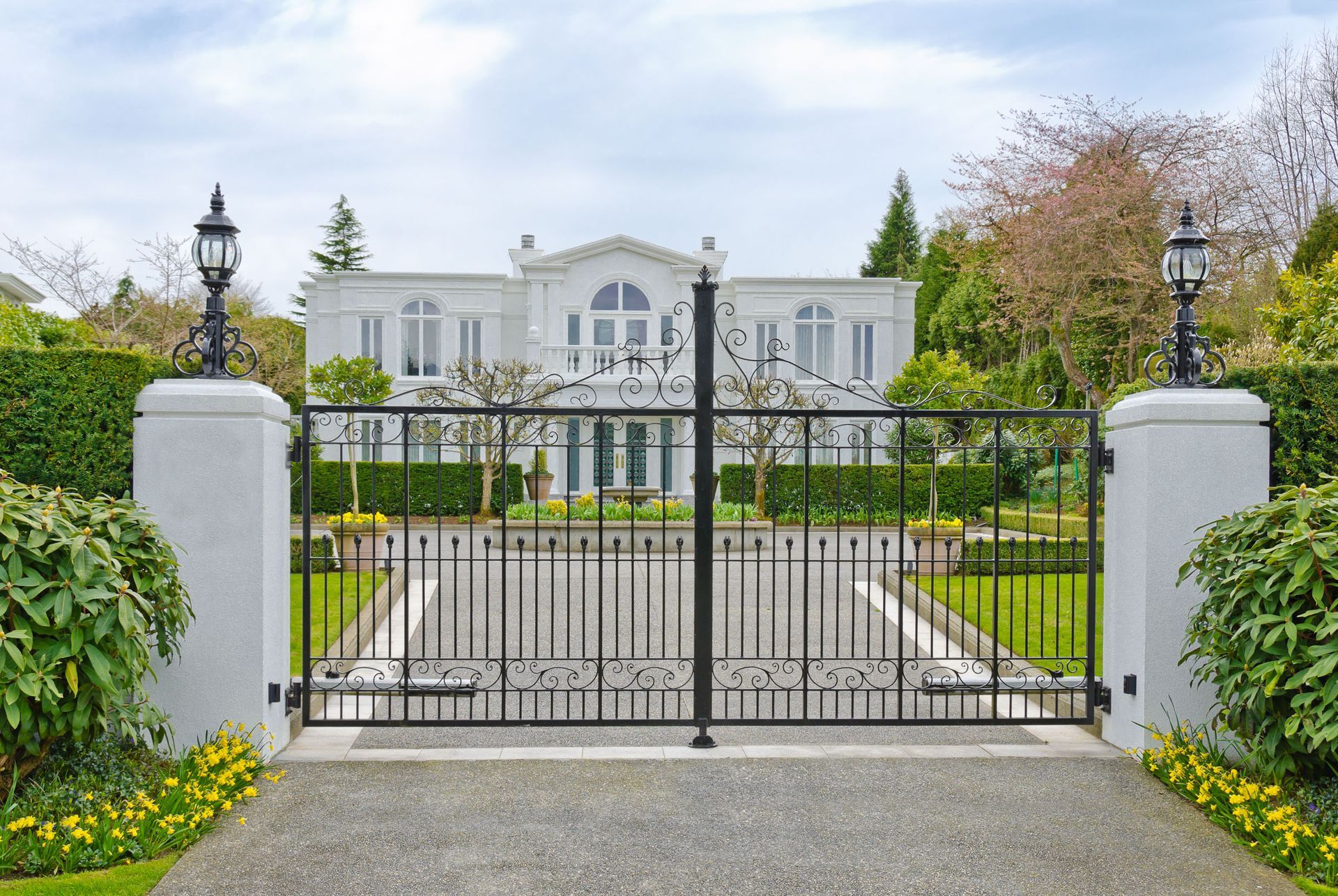 Gated driveway leading to a large white mansion with manicured gardens and stone pillars