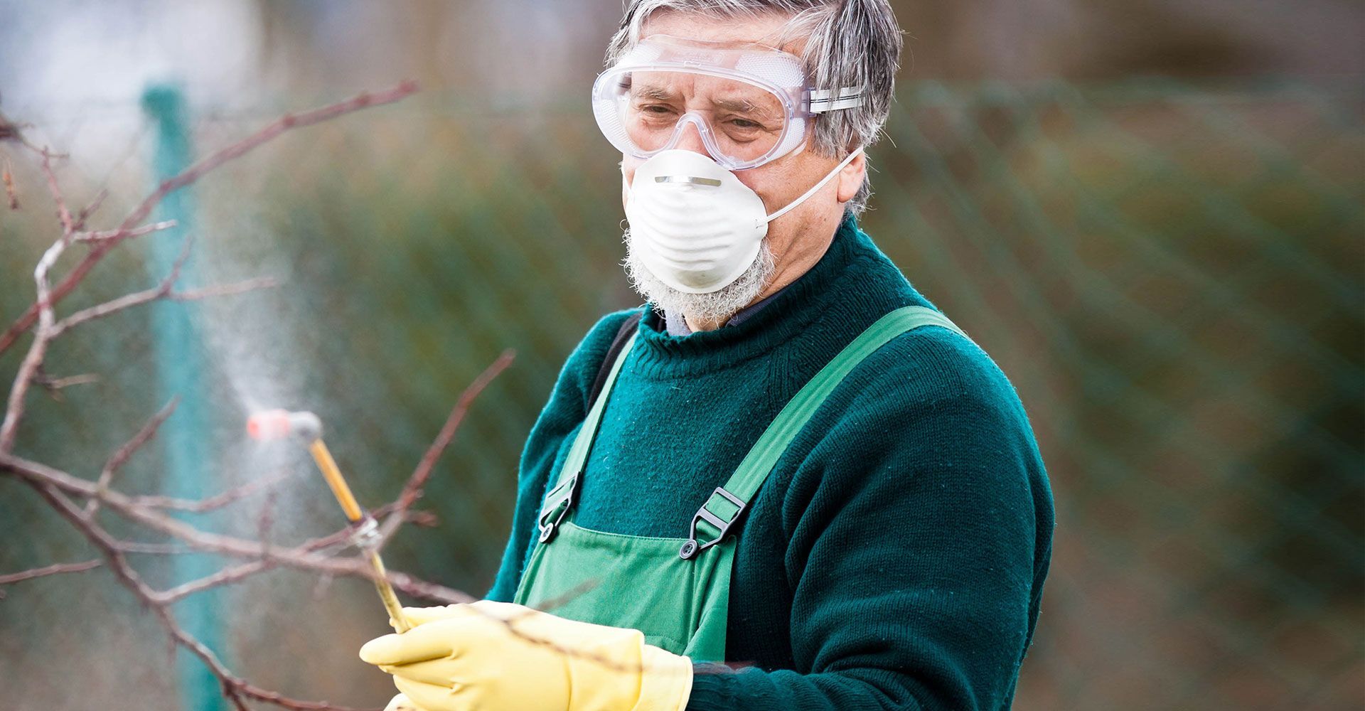 Person in protective gear pruning a tree branch outdoors, wearing goggles, mask, green sweater, and yellow gloves
