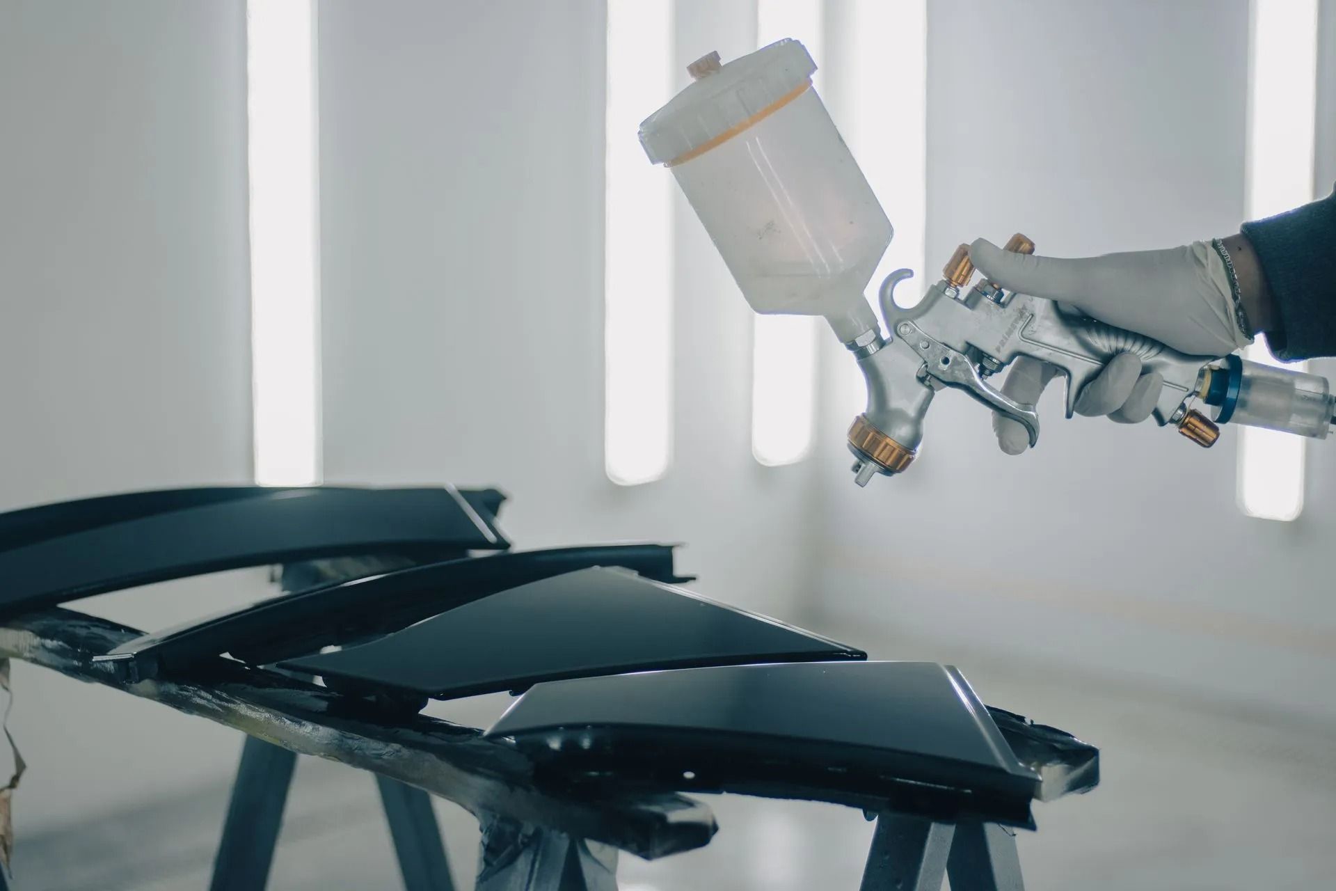 Person spraying paint on car parts with a spray gun in a brightly lit workshop.