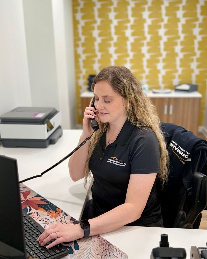 Woman with curly hair on phone at a desk, typing on a keyboard. Yellow patterned wall in the background.