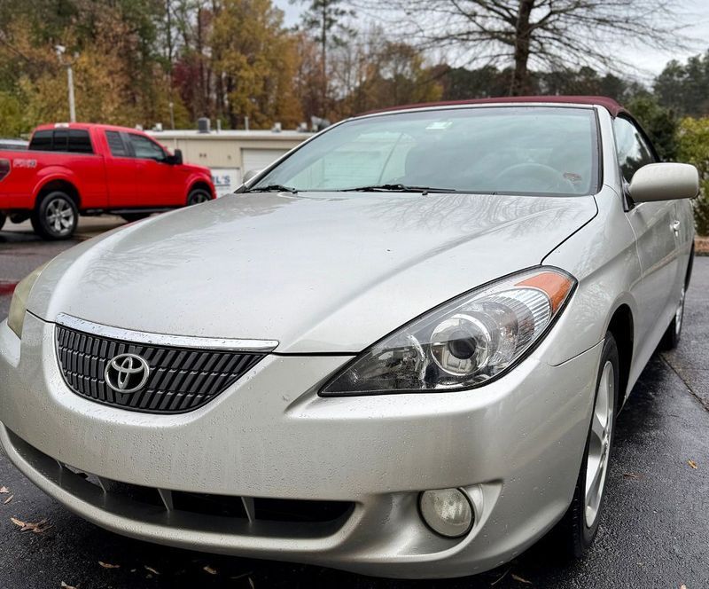 Silver Toyota Solara convertible parked on wet pavement, red truck in background.