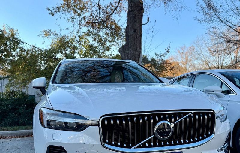 White Volvo XC60 SUV parked outside, front view. Chrome grill, tree in background, blue sky.