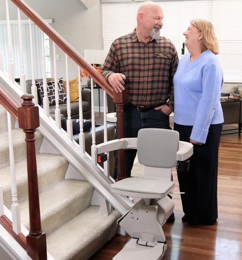 Stair lift beside a staircase, with a man and woman standing nearby in a home.