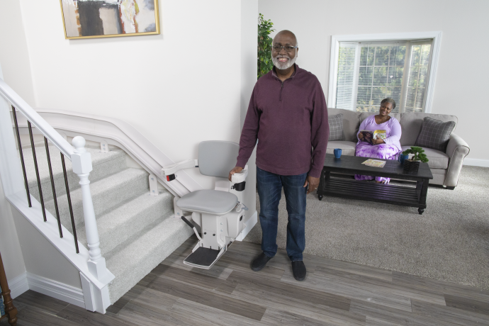 Man standing beside a stairlift in a bright living room, with a woman seated on a couch in the background.