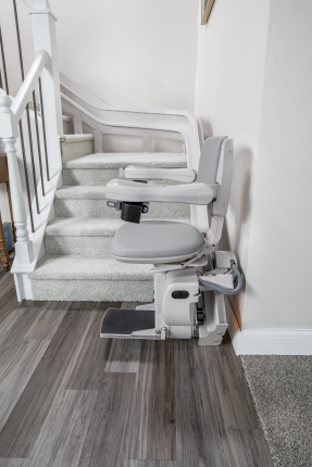 Indoor stair lift beside carpeted stairs in a white hallway, with gray swivel seat and footrest.