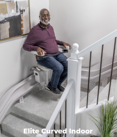 Man seated on a curved indoor stair lift, smiling in a stairwell with white railings and gray carpet.