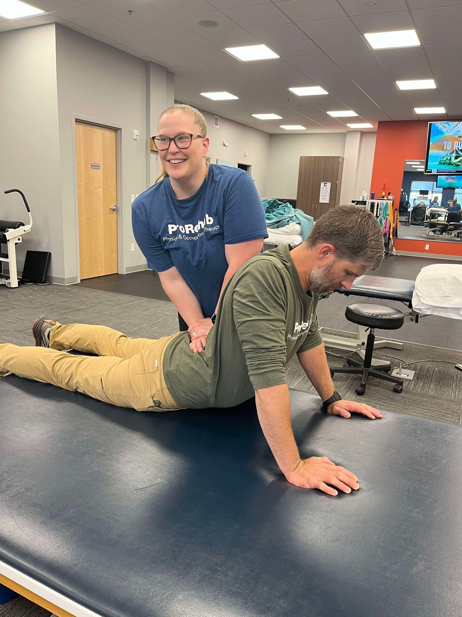 Therapist assists patient with back extension on a therapy table. Both are in a physical therapy clinic.