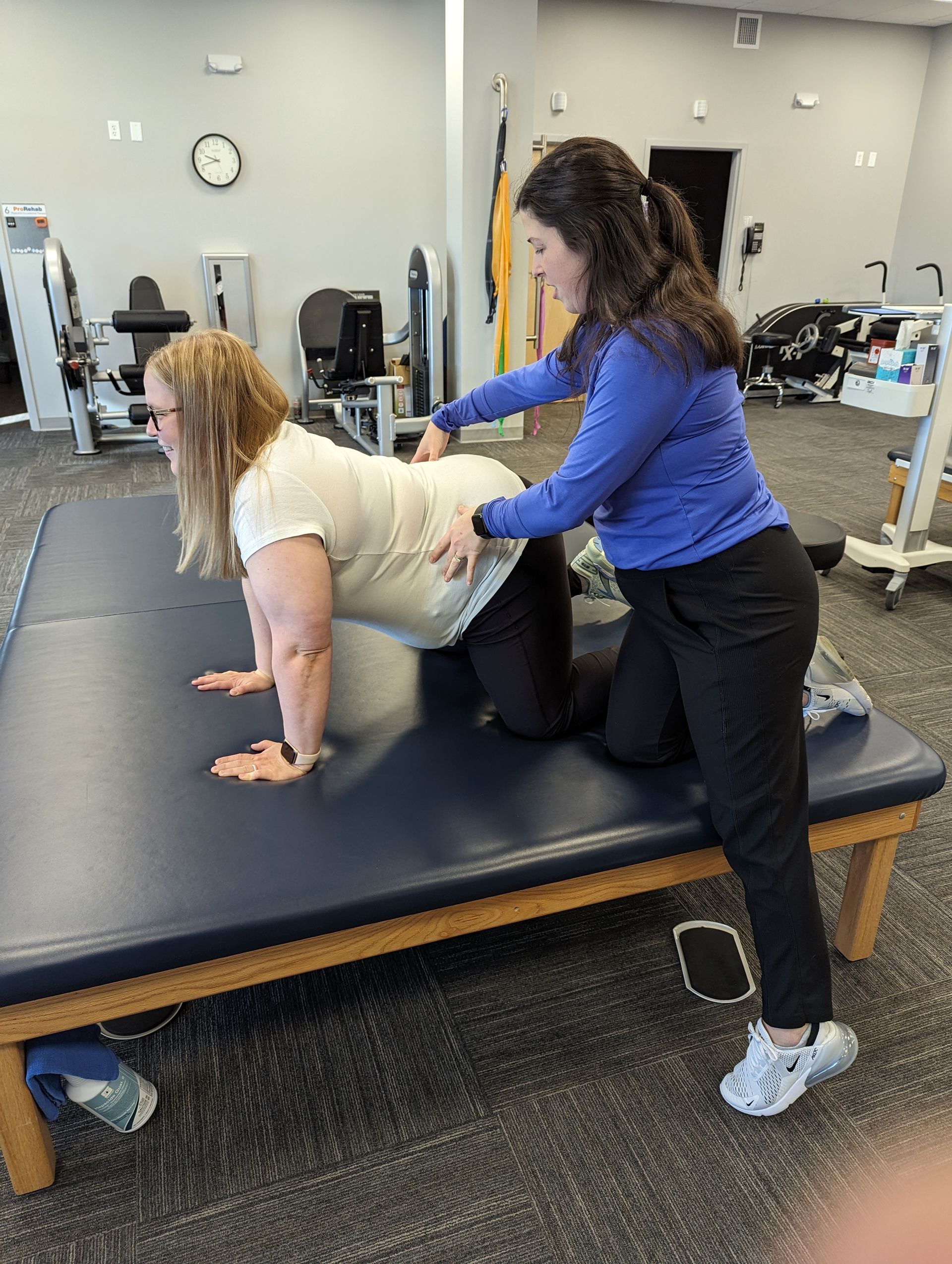 A therapist assists a person on hands and knees on a therapy table, hands on their back.
