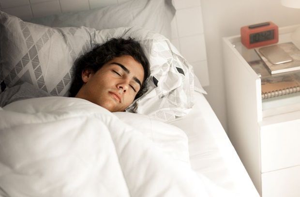 Young person asleep in bed, white bedding, nightstand with clock.