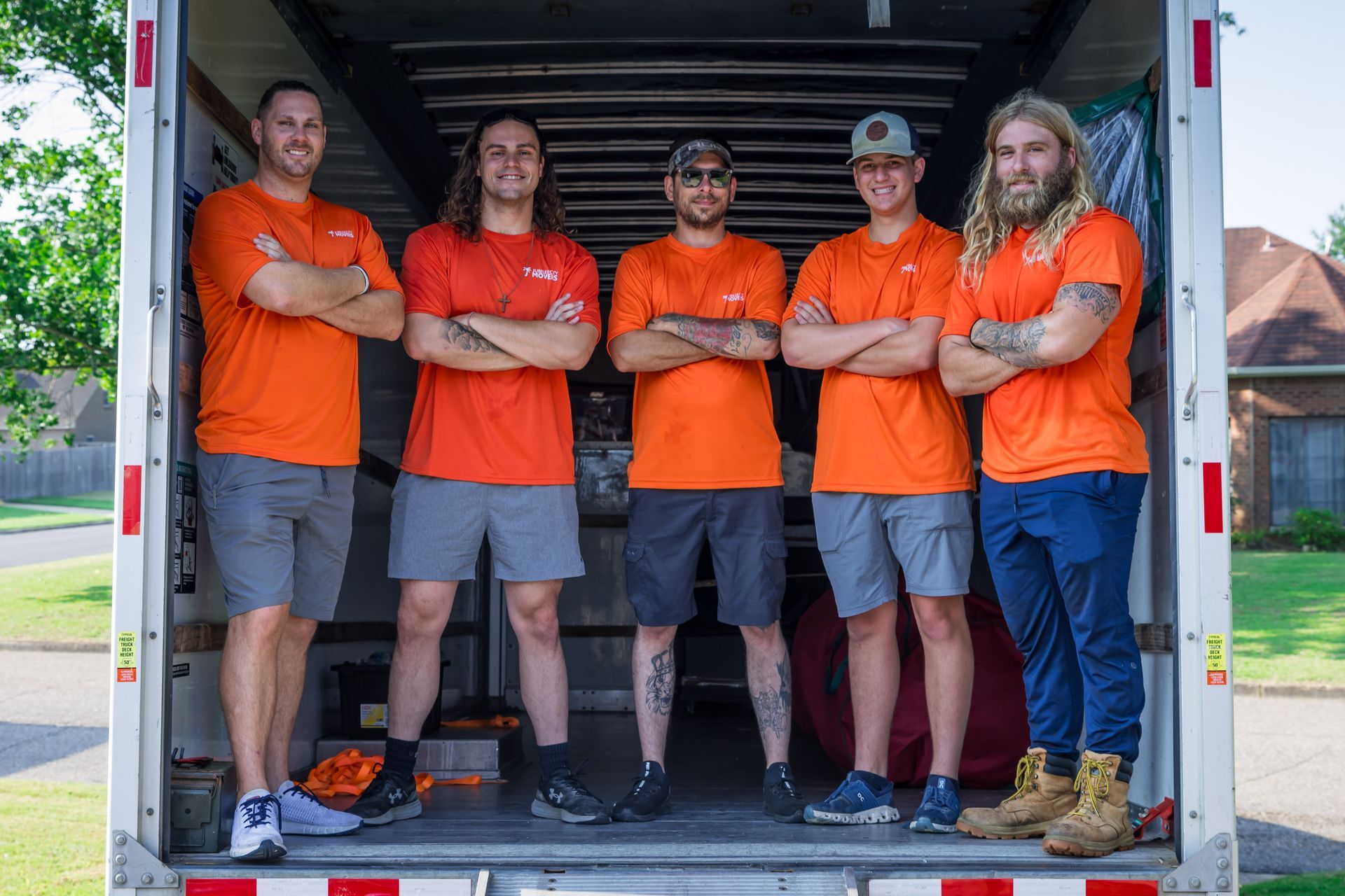 Five people wearing matching orange shirts stand with their arms crossed inside the back of an empty moving truck.