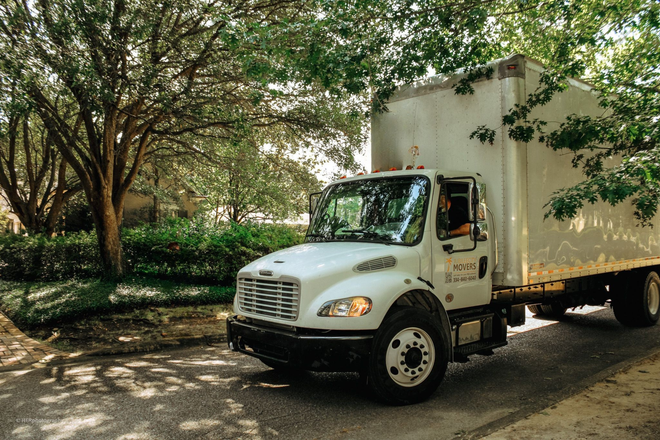 A white box truck driving on a tree-lined residential street during the day.