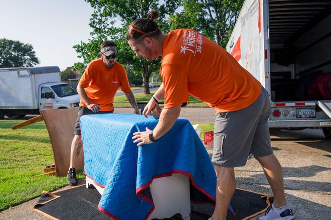 Two movers in orange shirts cover a piece of furniture with a blue moving blanket near an open moving truck.
