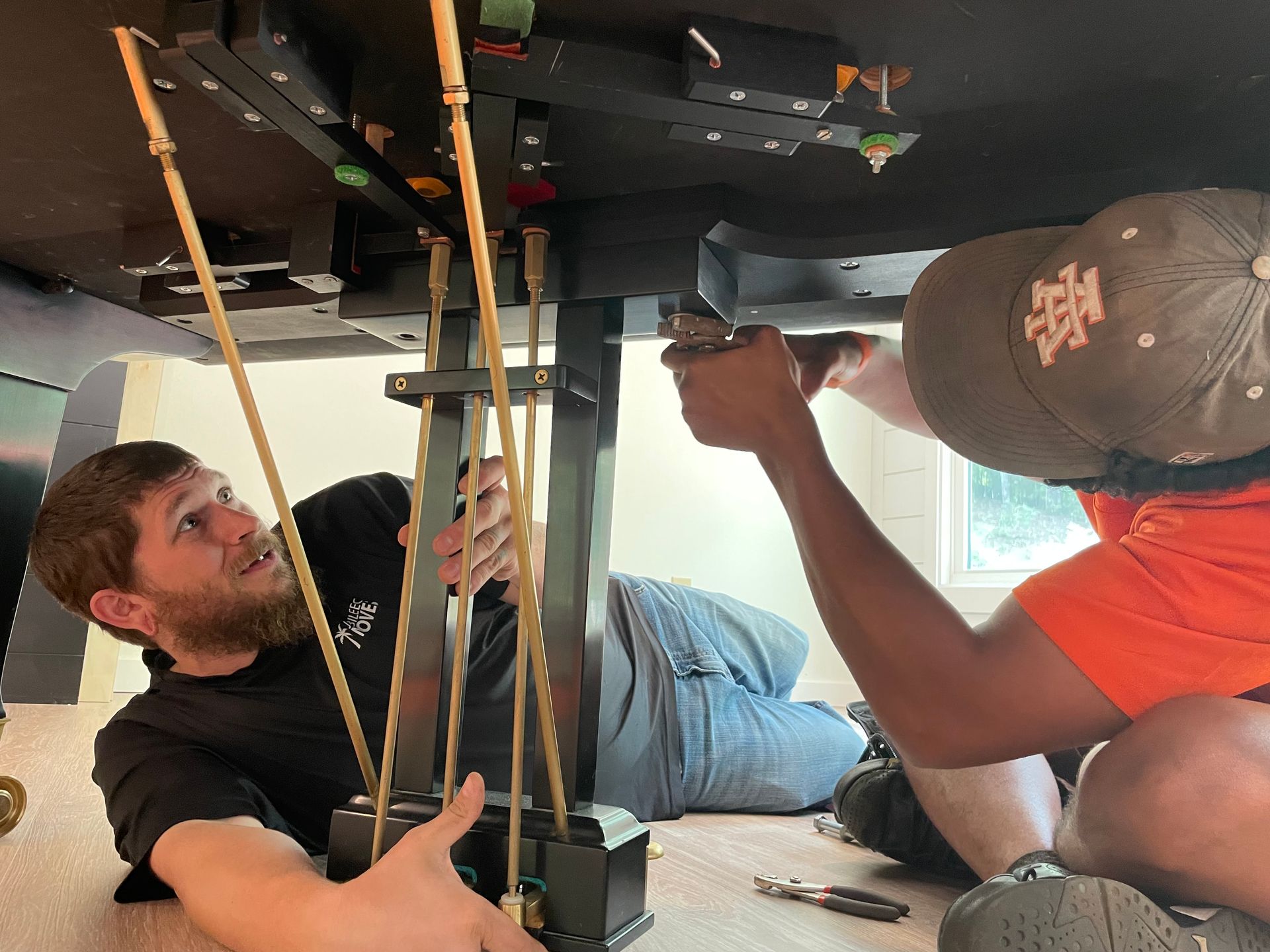 Two people installing a black structure, using tools, under a dark surface, one wearing a hat.