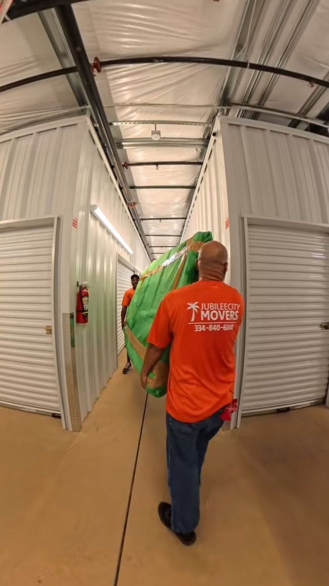 Two people are carrying a large green object down a hallway in a storage unit. One person wears an orange shirt.