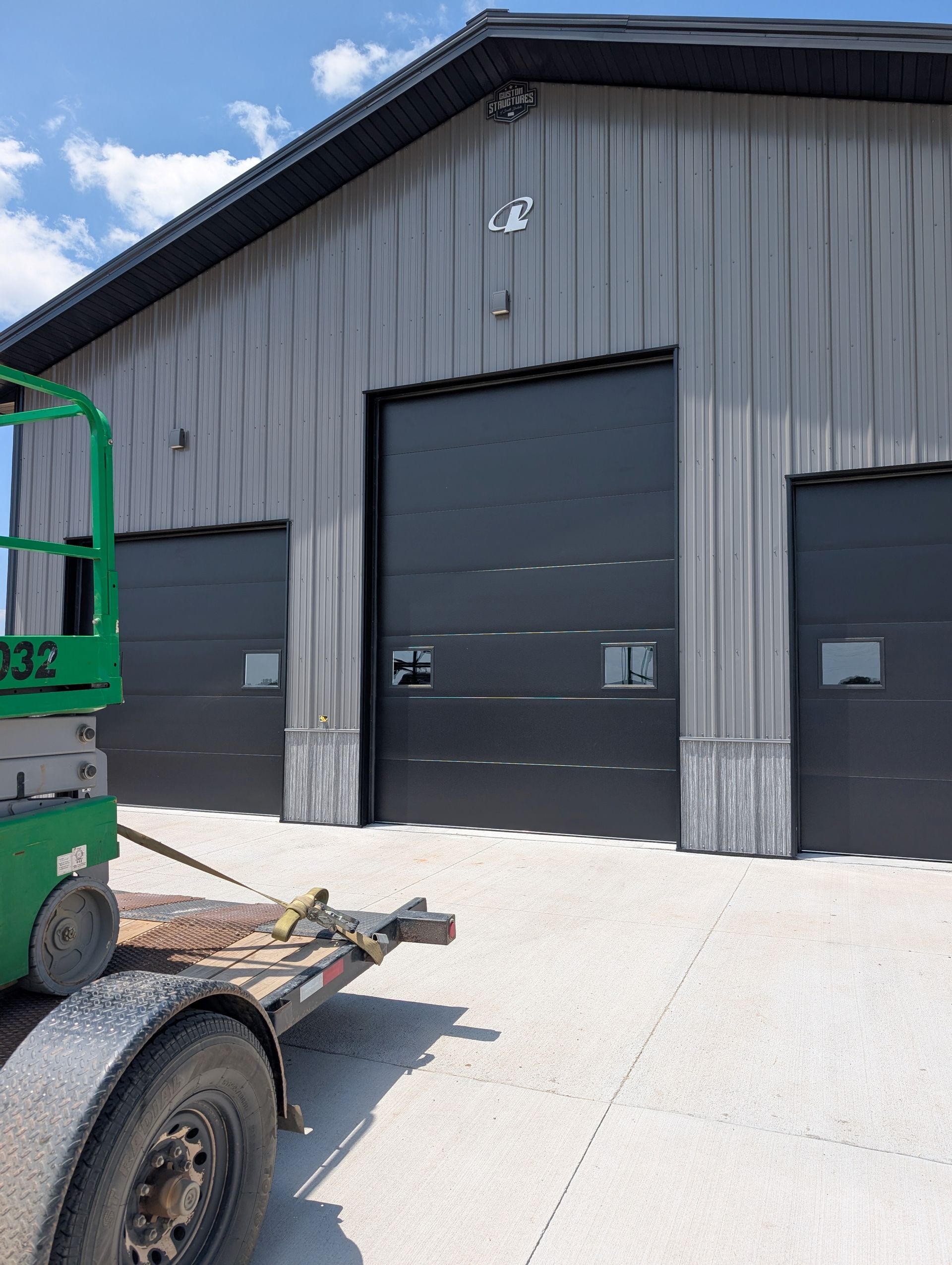 Gray industrial warehouse with two large black garage doors and a green lift at left