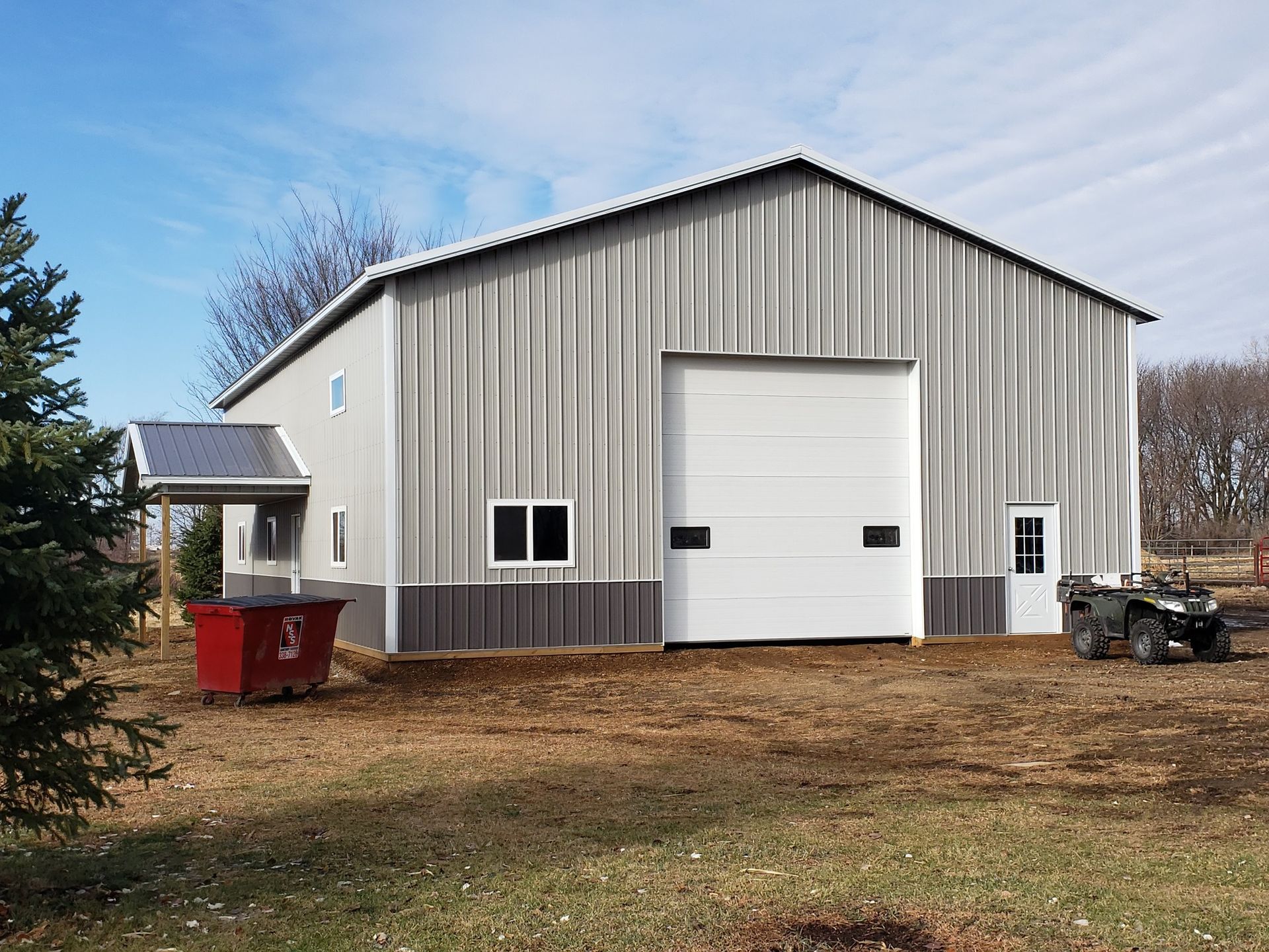 Large gray metal shed with white garage door and small side entrance, red dumpster, and an ATV.