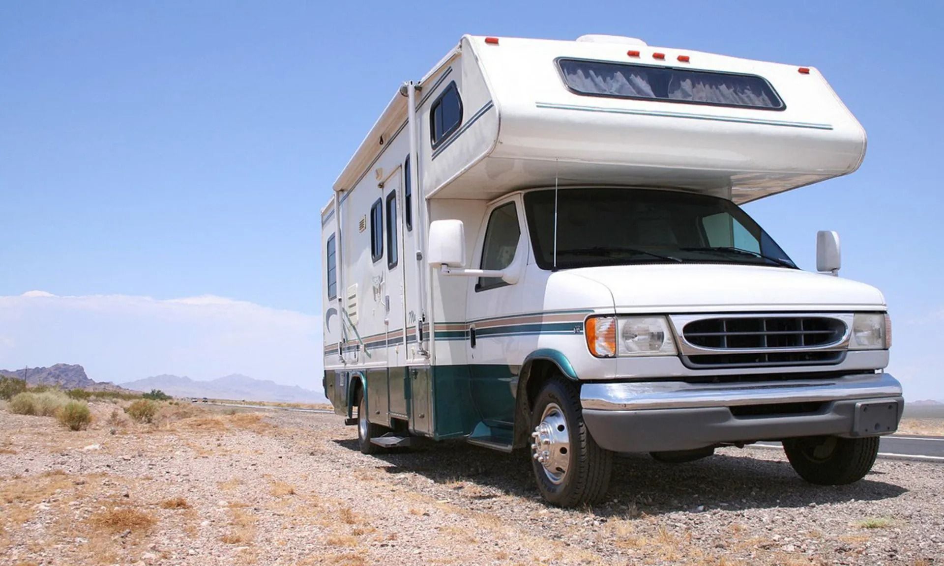 White and teal RV parked on a gravel road, with a blue sky background and desert landscape.