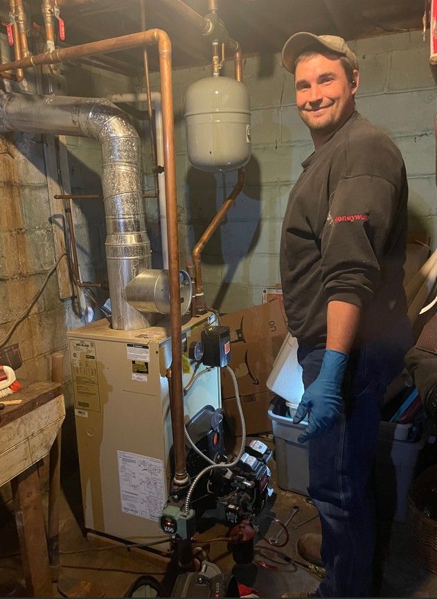 Man smiles next to furnace in basement; copper pipes, silver tank, dark clothes, blue gloves.