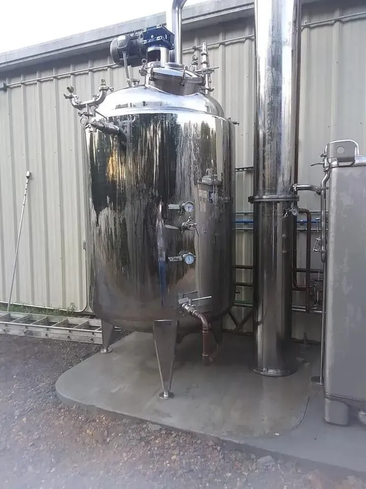 Stainless steel industrial tank with pipes and equipment. Outdoors against a building.